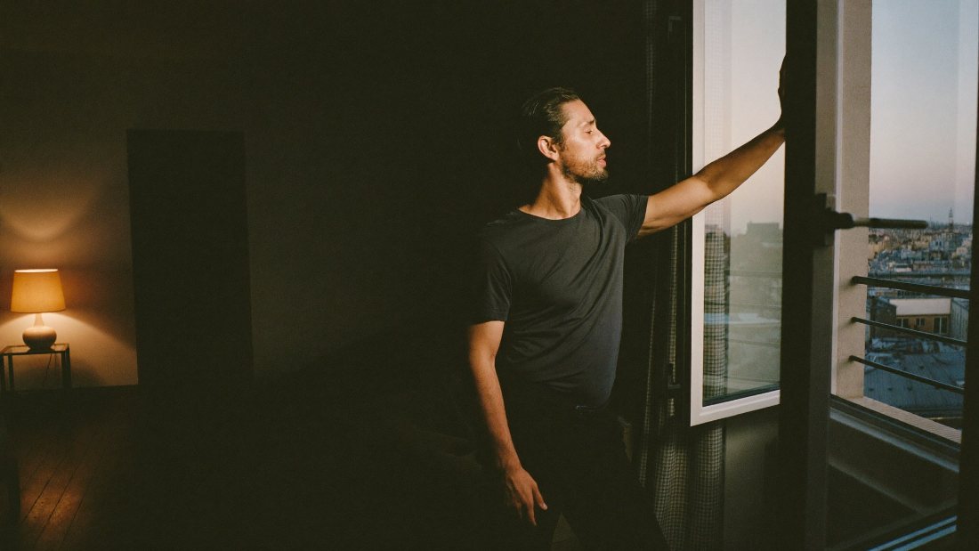 A man standing at an open window in a lamp-lit apartment in the evening.